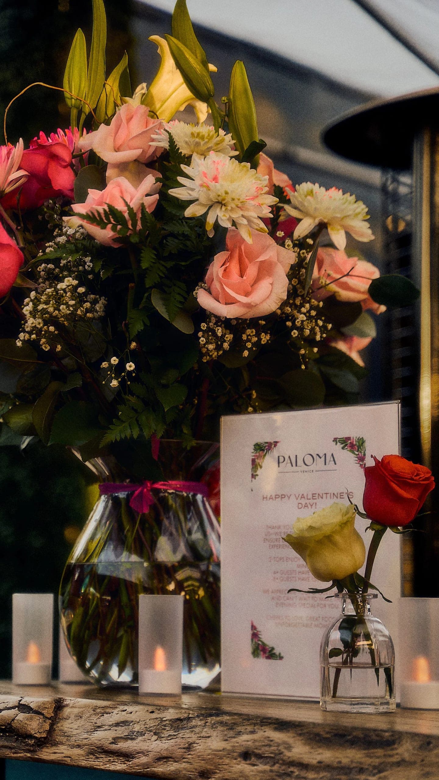 A Paloma menu card propped between a vase of roses and lilies and a single red rose