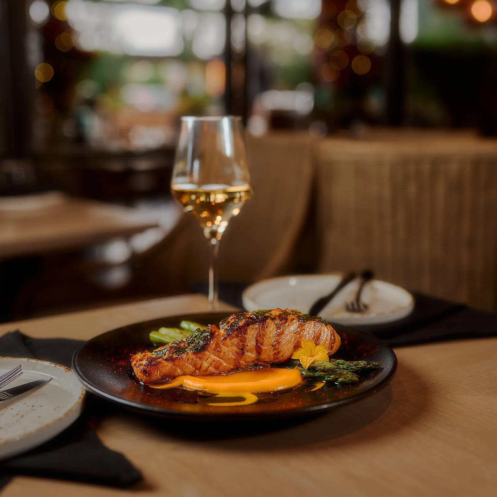 A plated fish course beside a glass of white wine on a linen-topped table