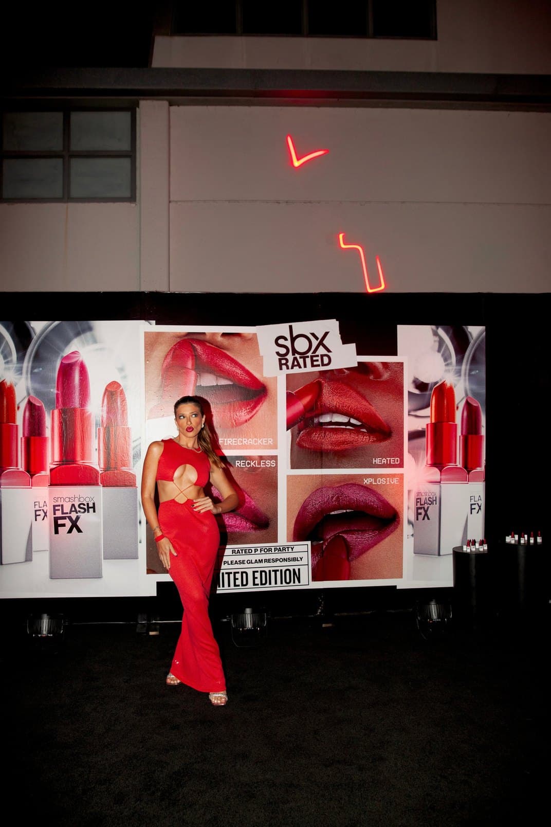 A guest in red posing in front of the Smashbox wall of oversized lipstick posters.