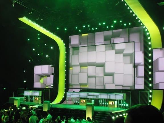 A stage framed in green light, with a wall of white cubes behind it.
