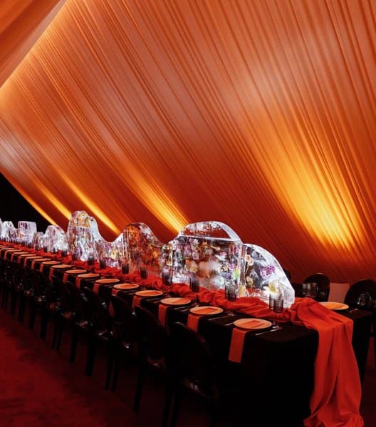 A long dinner table beneath sweeping orange draping, set with glass centrepieces and red runners.