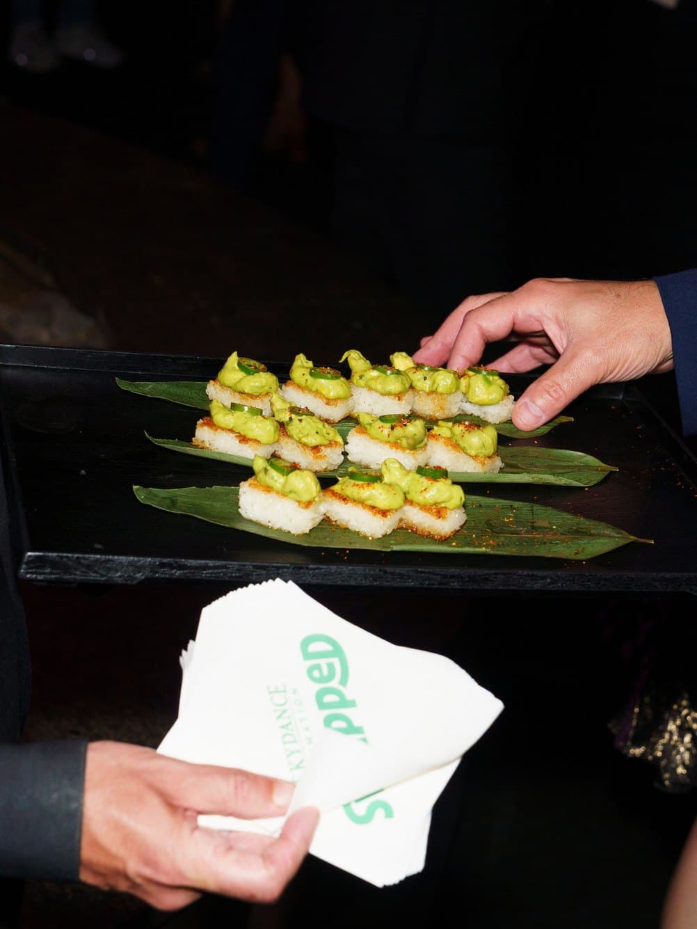 A tray of rice canapés topped with avocado, passed to guests with a cocktail napkin.