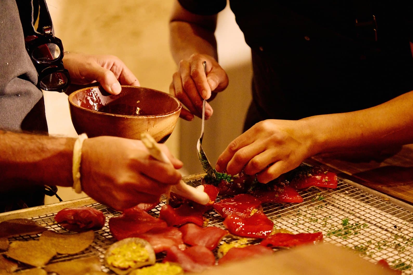 Chefs’ hands finishing charred red peppers on a grill rack, a bowl of salsa held over them.