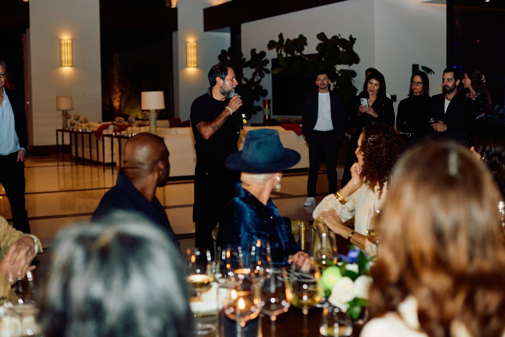 A host speaking into a microphone to guests seated at a candlelit dinner.