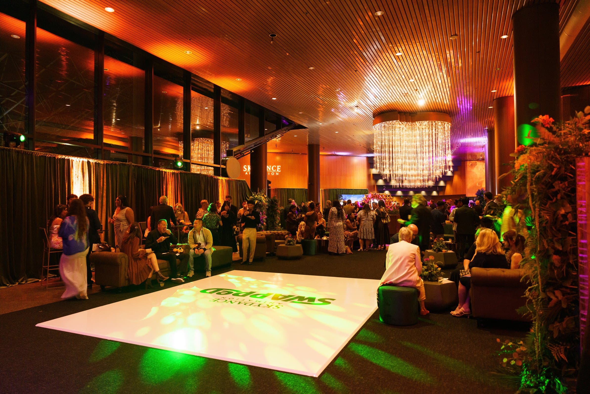 A lit dance floor at the centre of a lounge under a crystal chandelier, guests gathered at low tables around it.