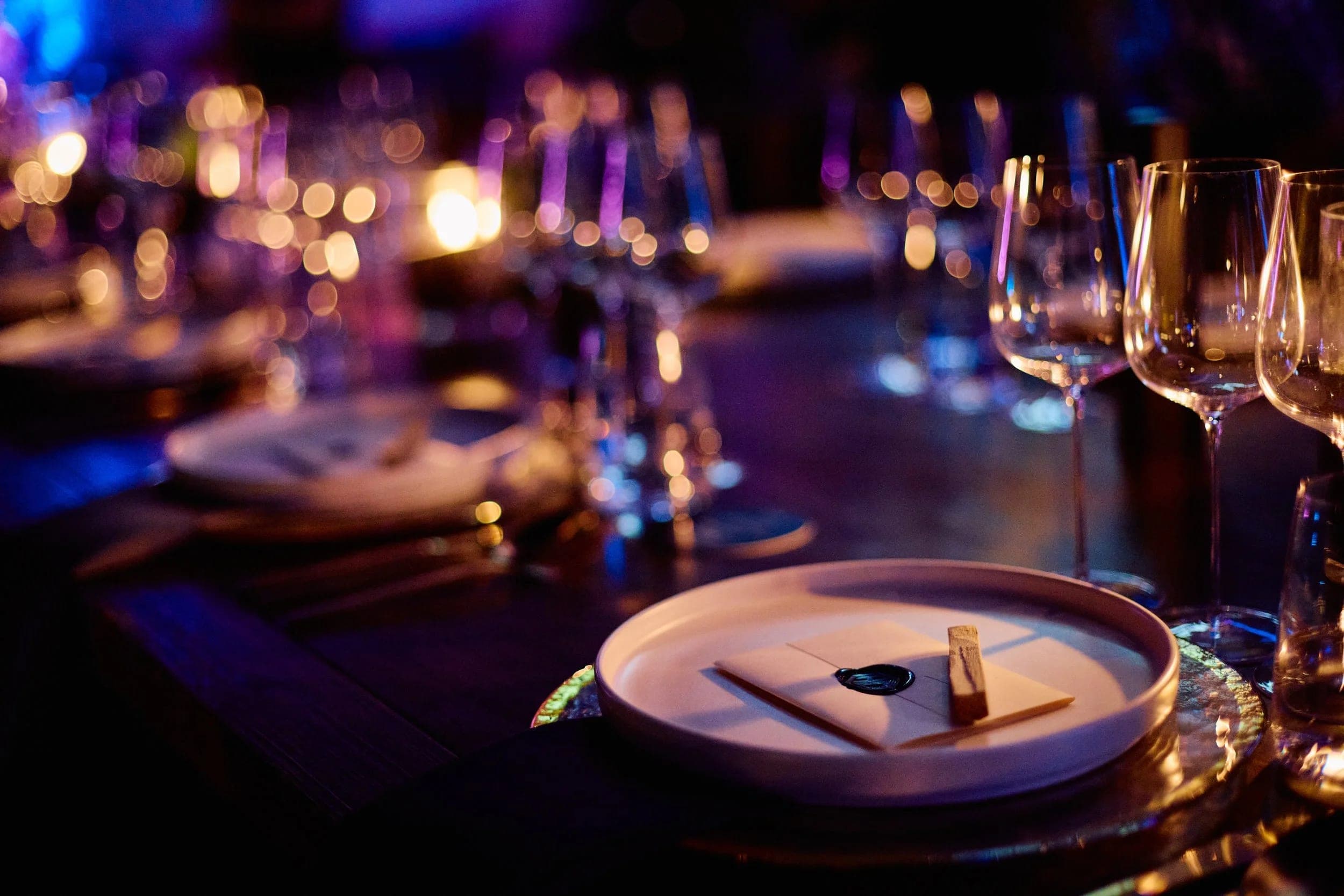 Candlelight and glassware blurred along a set dinner table, a place card resting on a plate