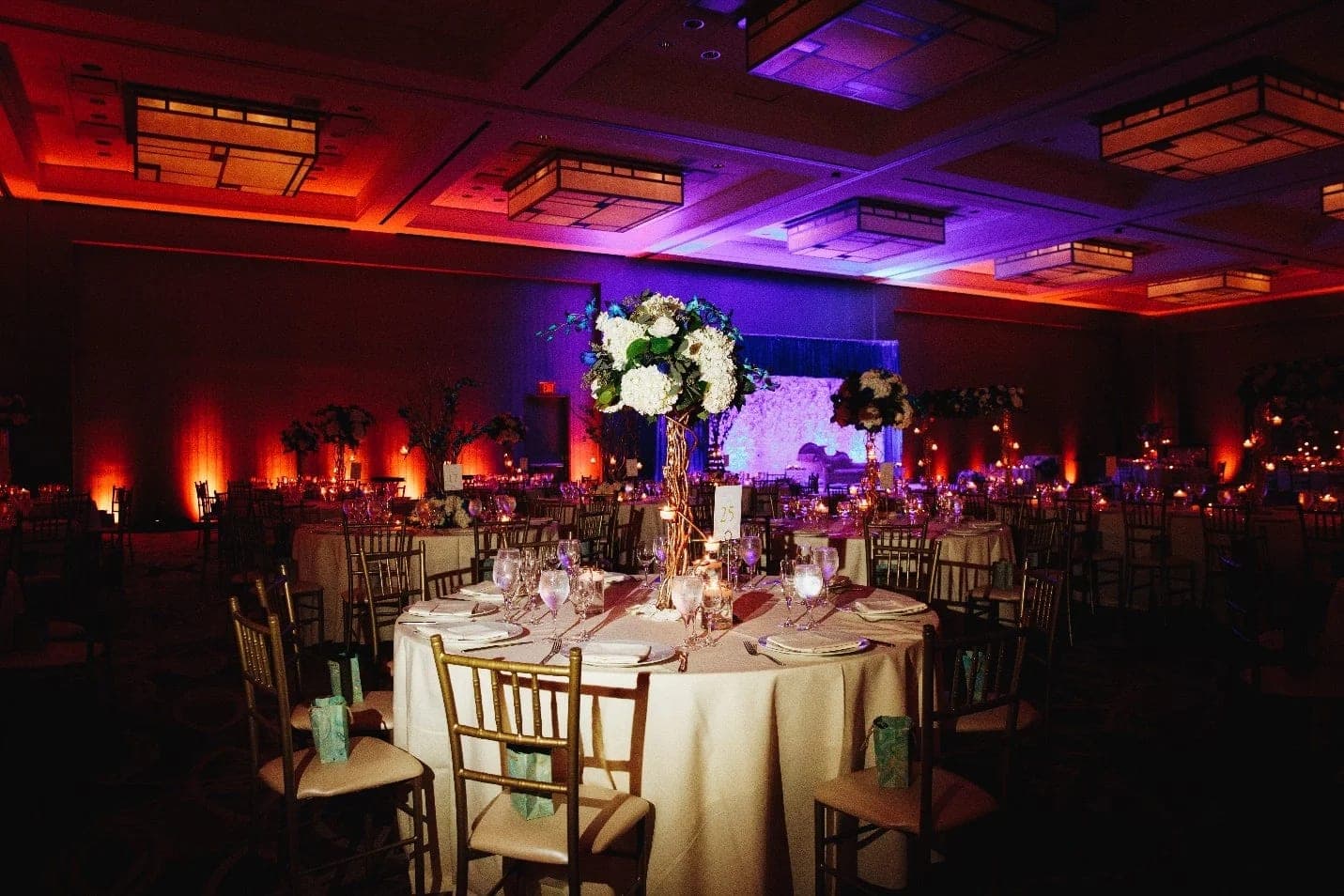 A ballroom dressed for dinner, round tables under amber and violet uplighting