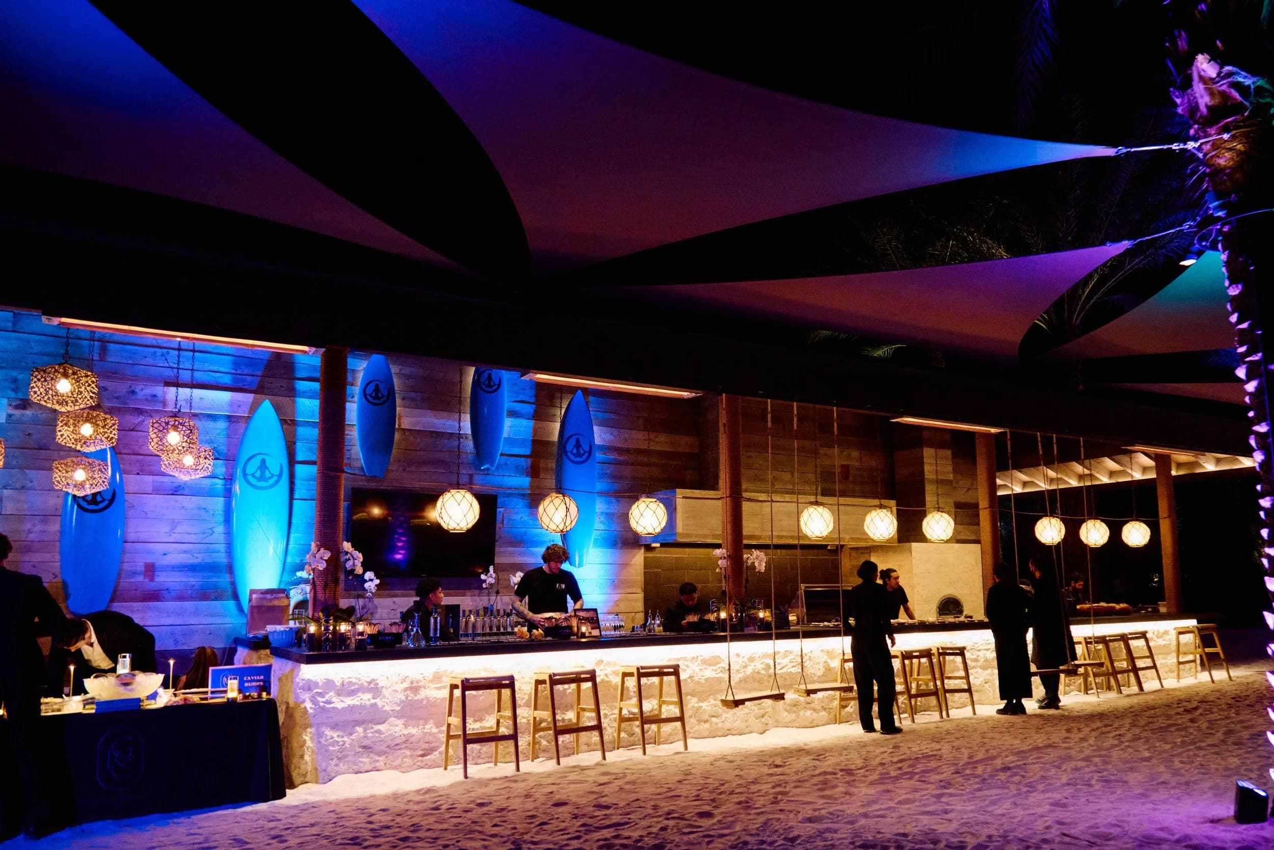 An outdoor bar beneath angular shade sails, lit blue and violet after dark
