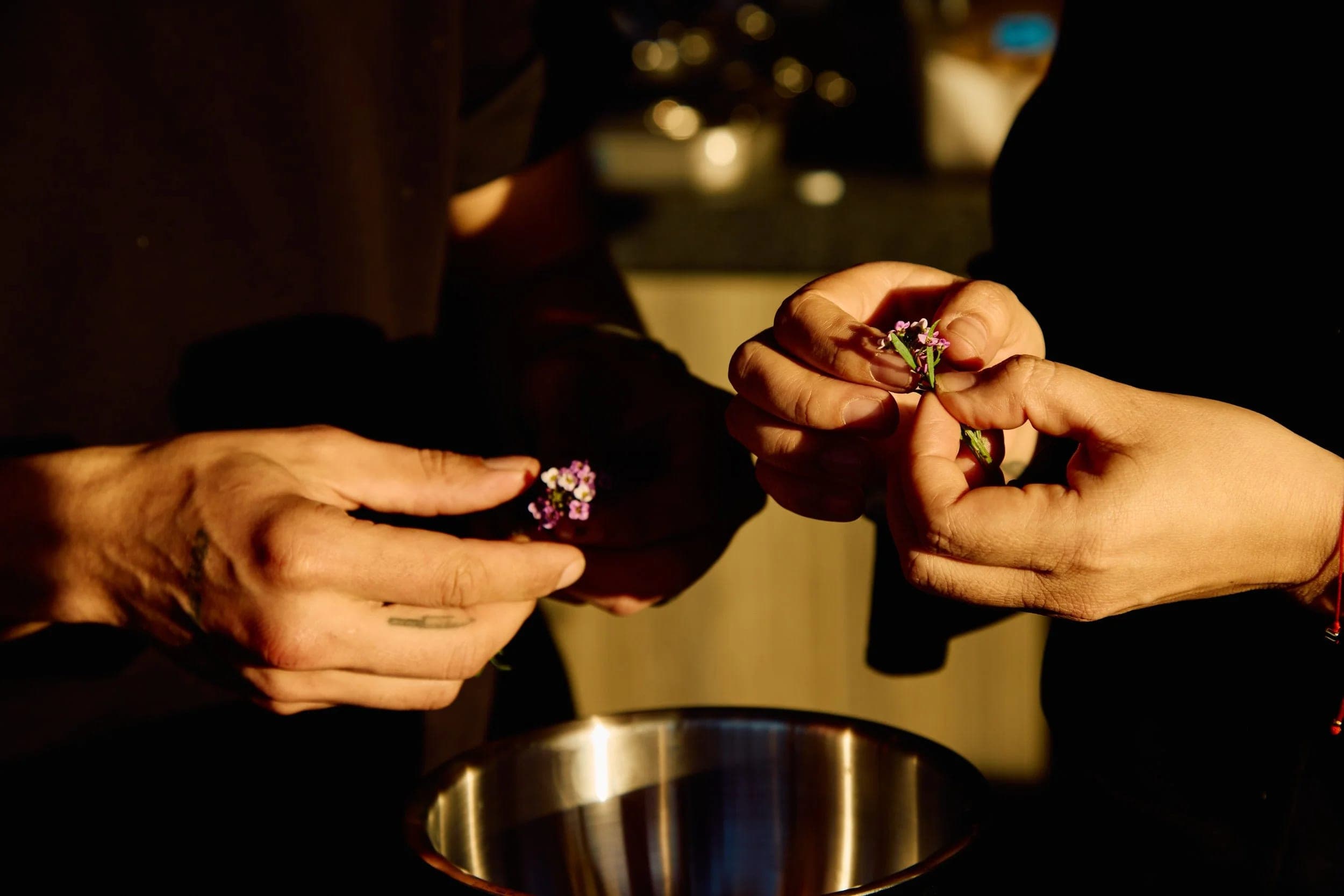 Two pairs of hands meeting over a brass bowl, passing a garnish between them