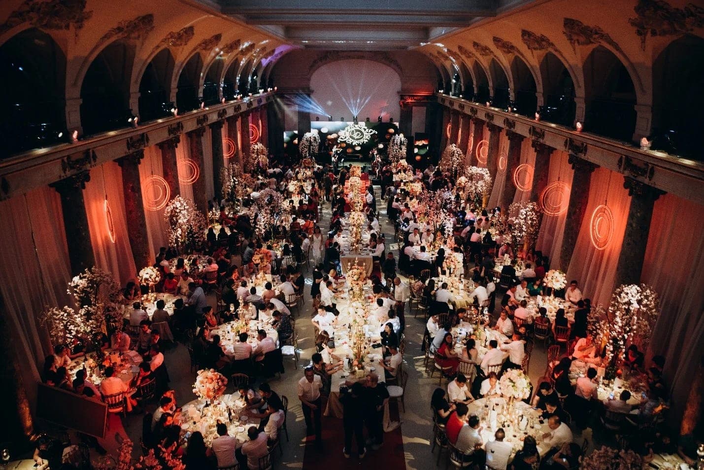 A vaulted hall filled with round tables and towering floral centrepieces, seen from the balcony
