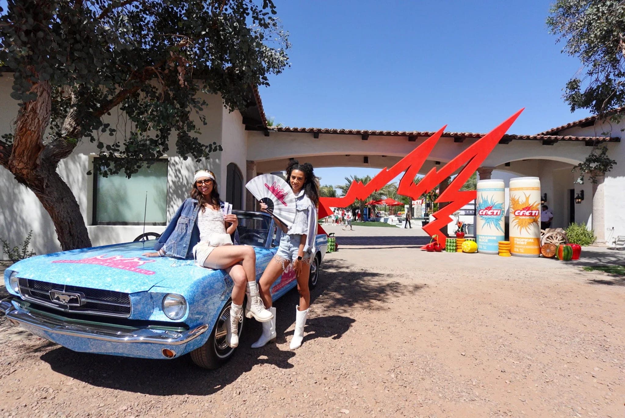 Two guests perched on a hand-painted vintage Mustang outside the Cacti pool party villa, red flags flying behind them