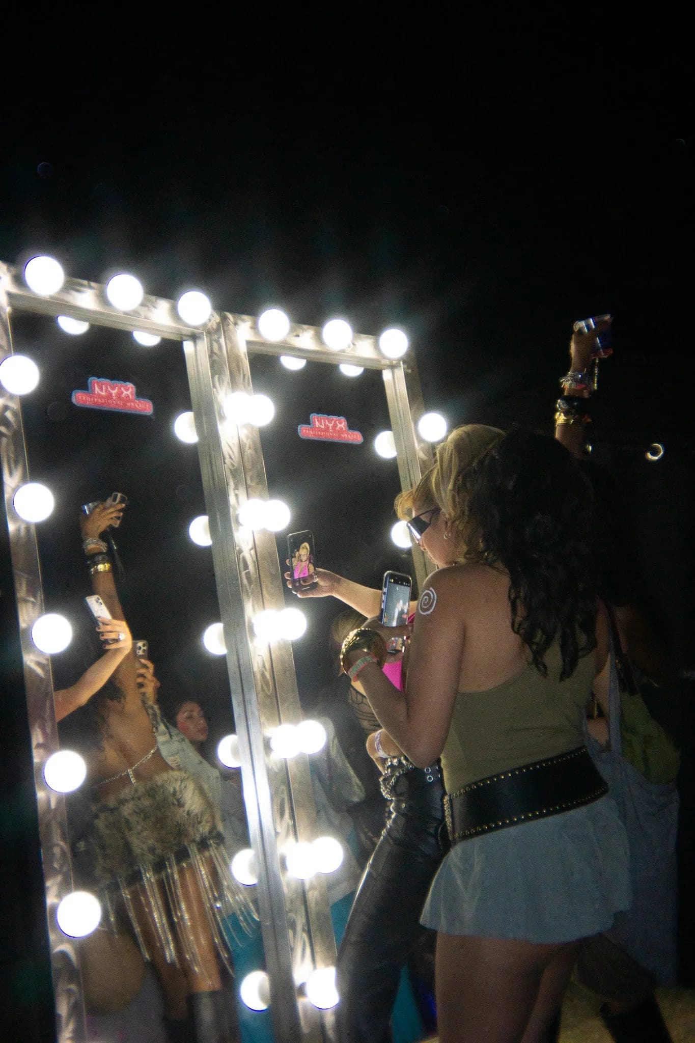 Guests gathered around a bulb-lit vanity mirror at Neon Carnival, drinks raised
