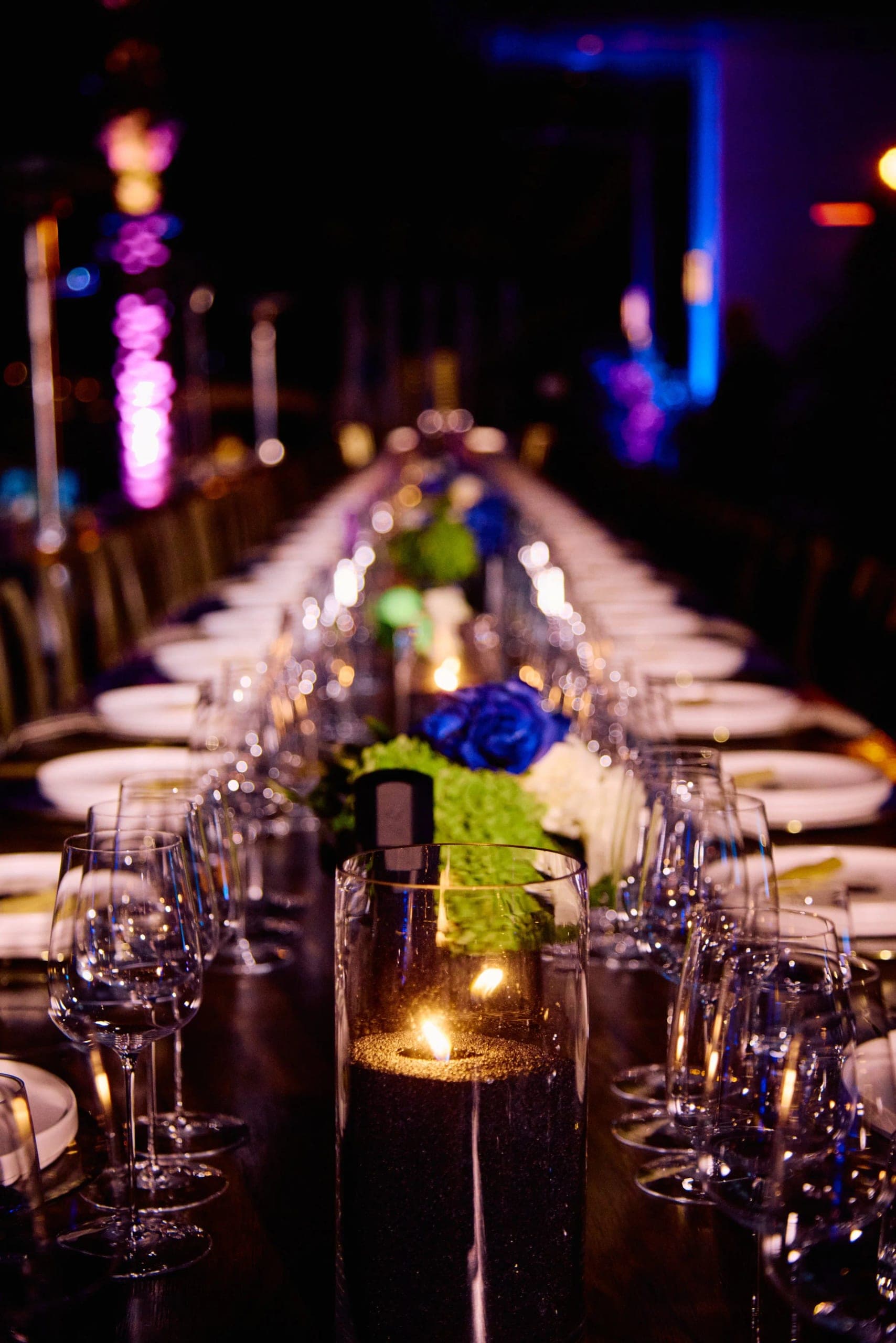 The banquet table after dark, a single candle and blue florals between rows of glassware receding into the distance