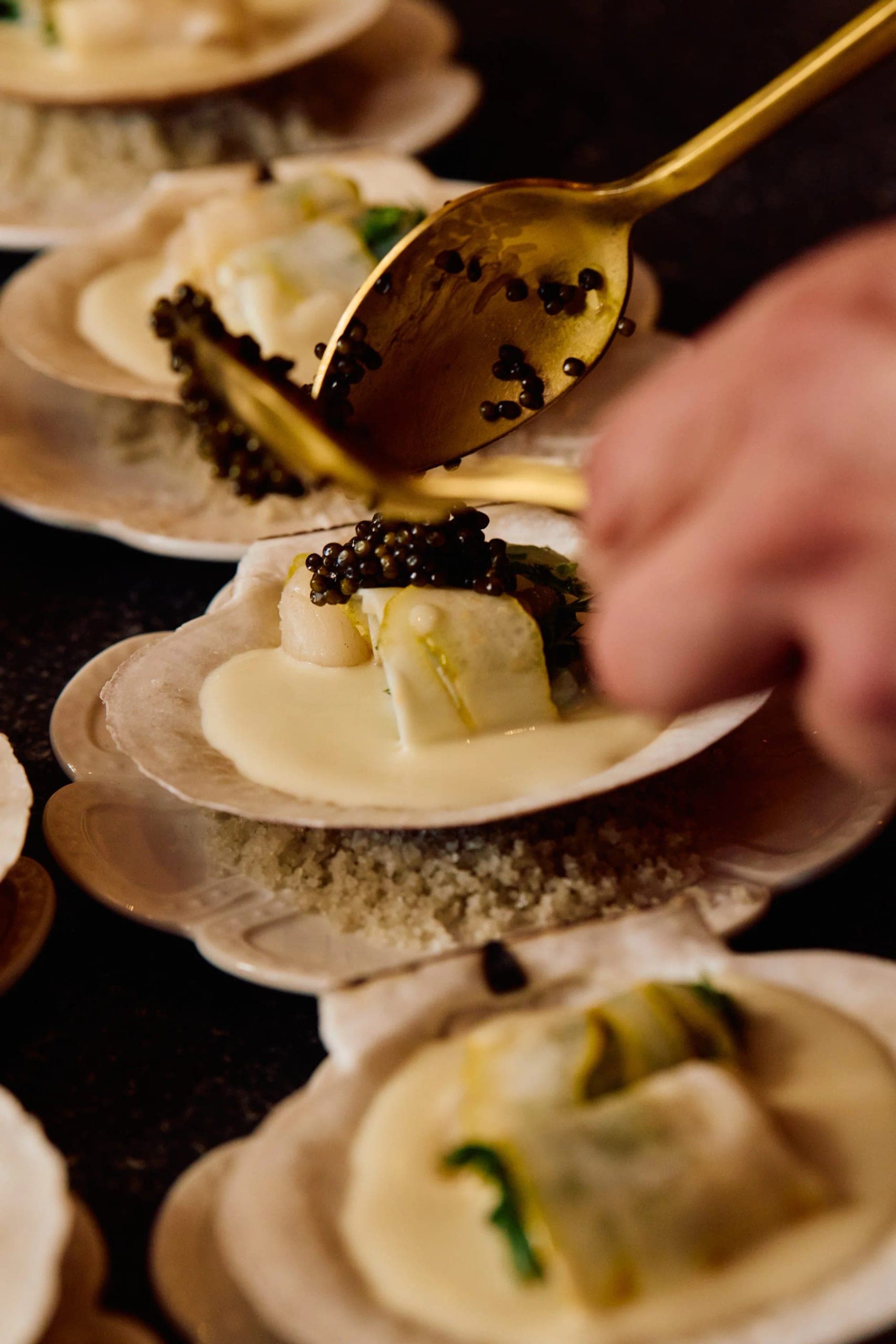 A gold spoon laying caviar onto a scallop shell, plated courses lined up behind it ready for service