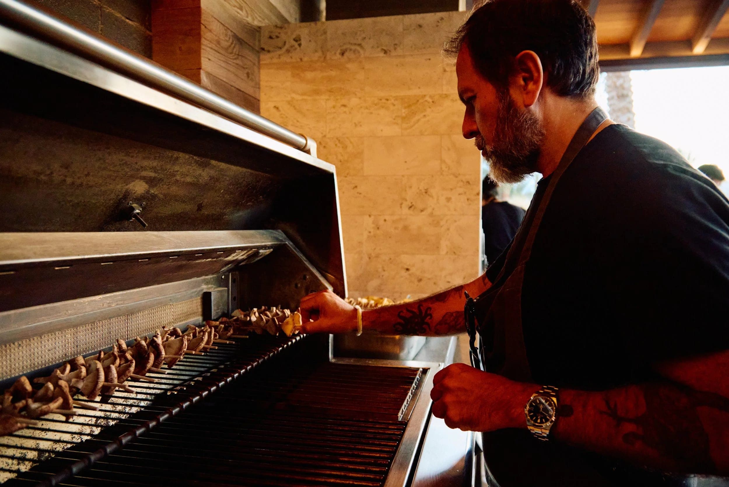 A chef lifting the hood of an open fire grill loaded with skewers, warm afternoon light across his arms