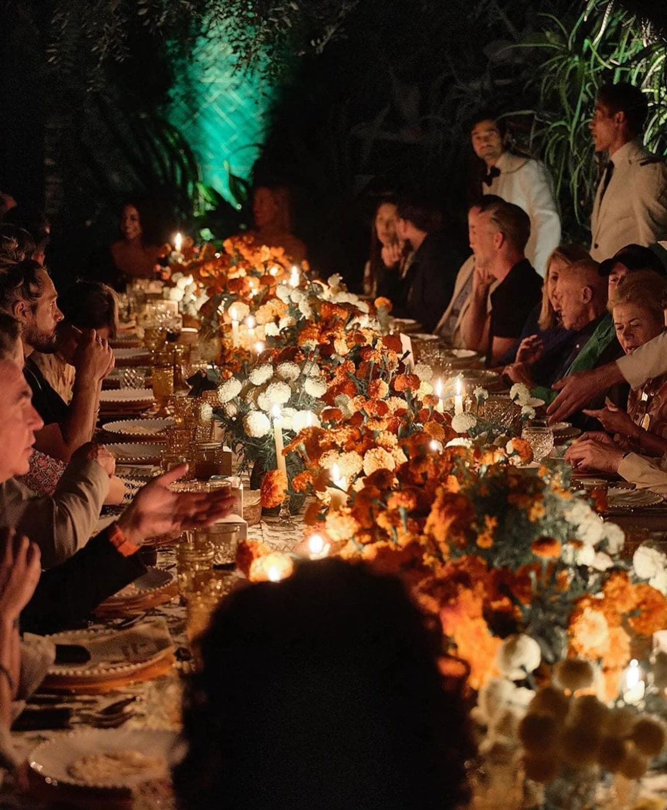 Guests seated the length of a candlelit table at night, flowers and hurricane lamps running down the centre