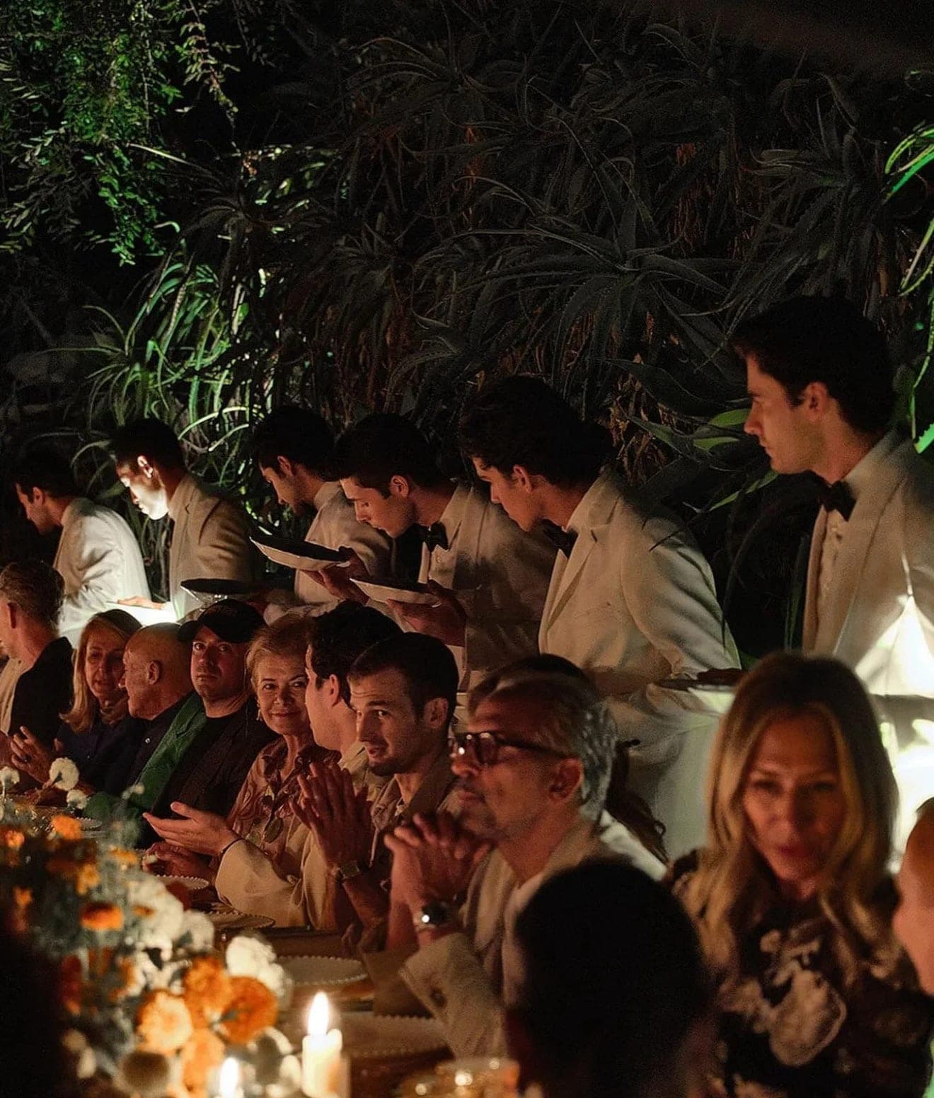 Guests in cream jackets leaning in over dinner as the course is served