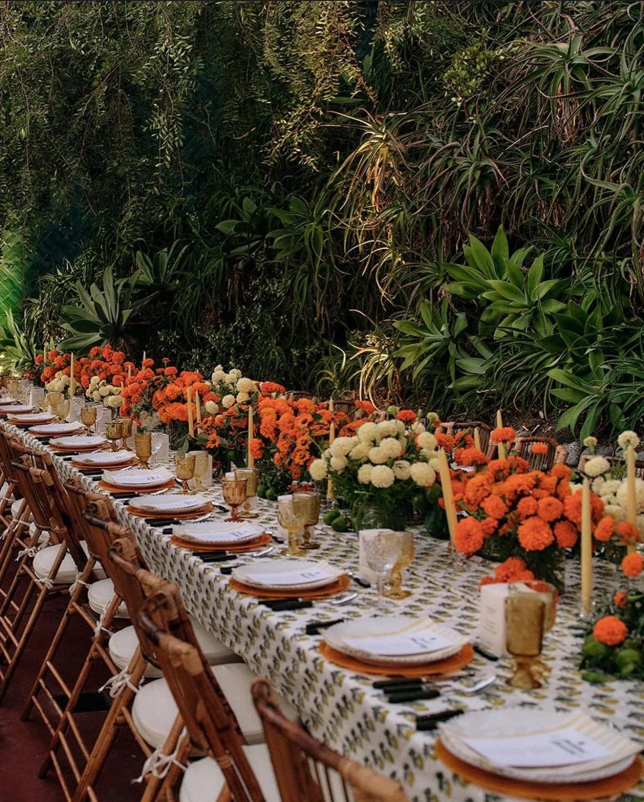 A long dinner table dressed with marigolds, cream tapers and printed linen, set among dense garden planting
