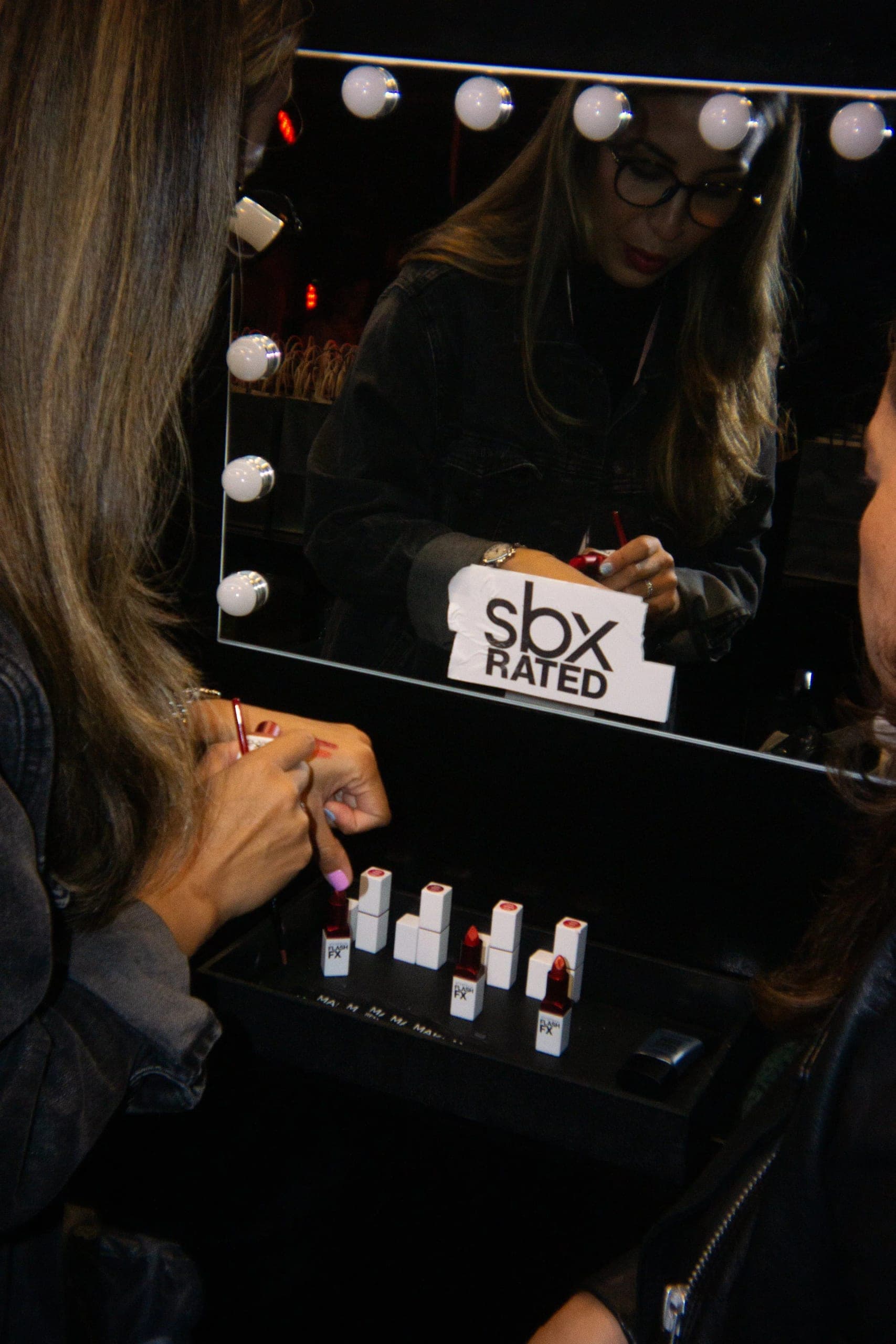 A makeup artist working at a bulb-lit vanity mirror with lipsticks laid out beneath it