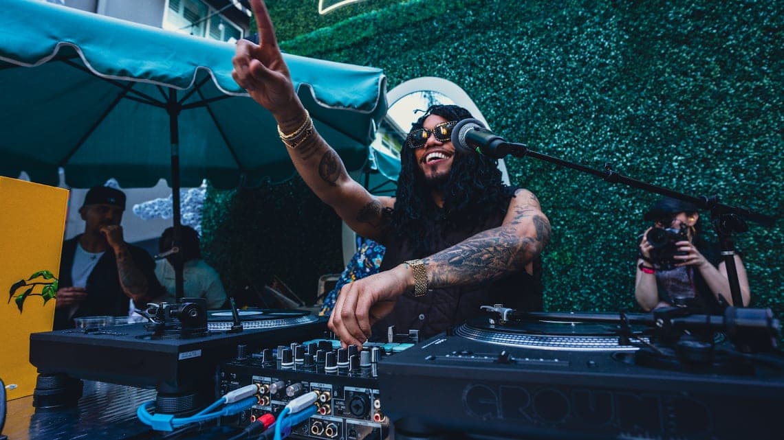 The DJ throwing an arm up behind the decks beneath a green parasol