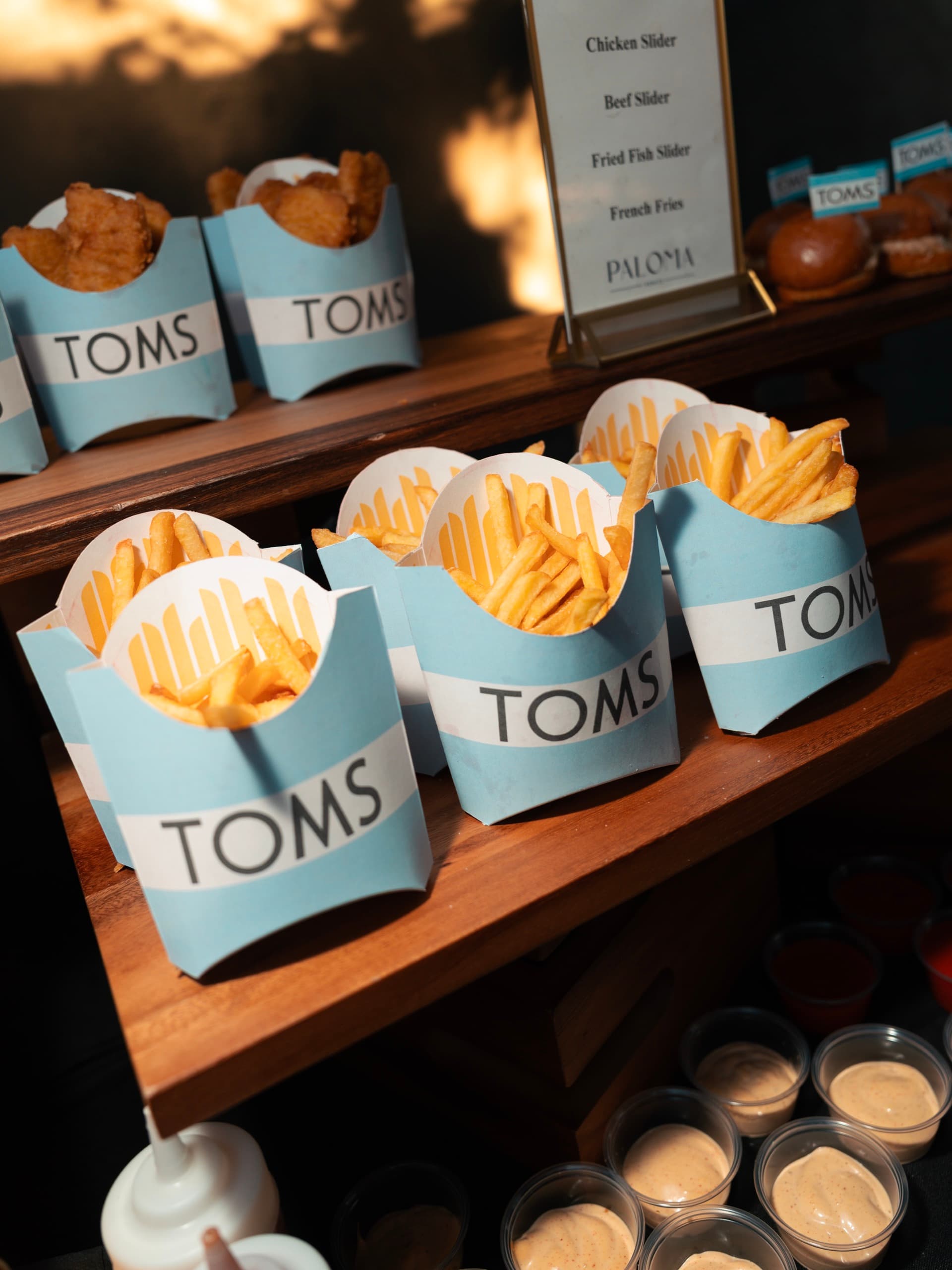TOMS-branded cartons of fries and chicken tenders on a wooden shelf beside the printed Paloma menu of sliders and fries.