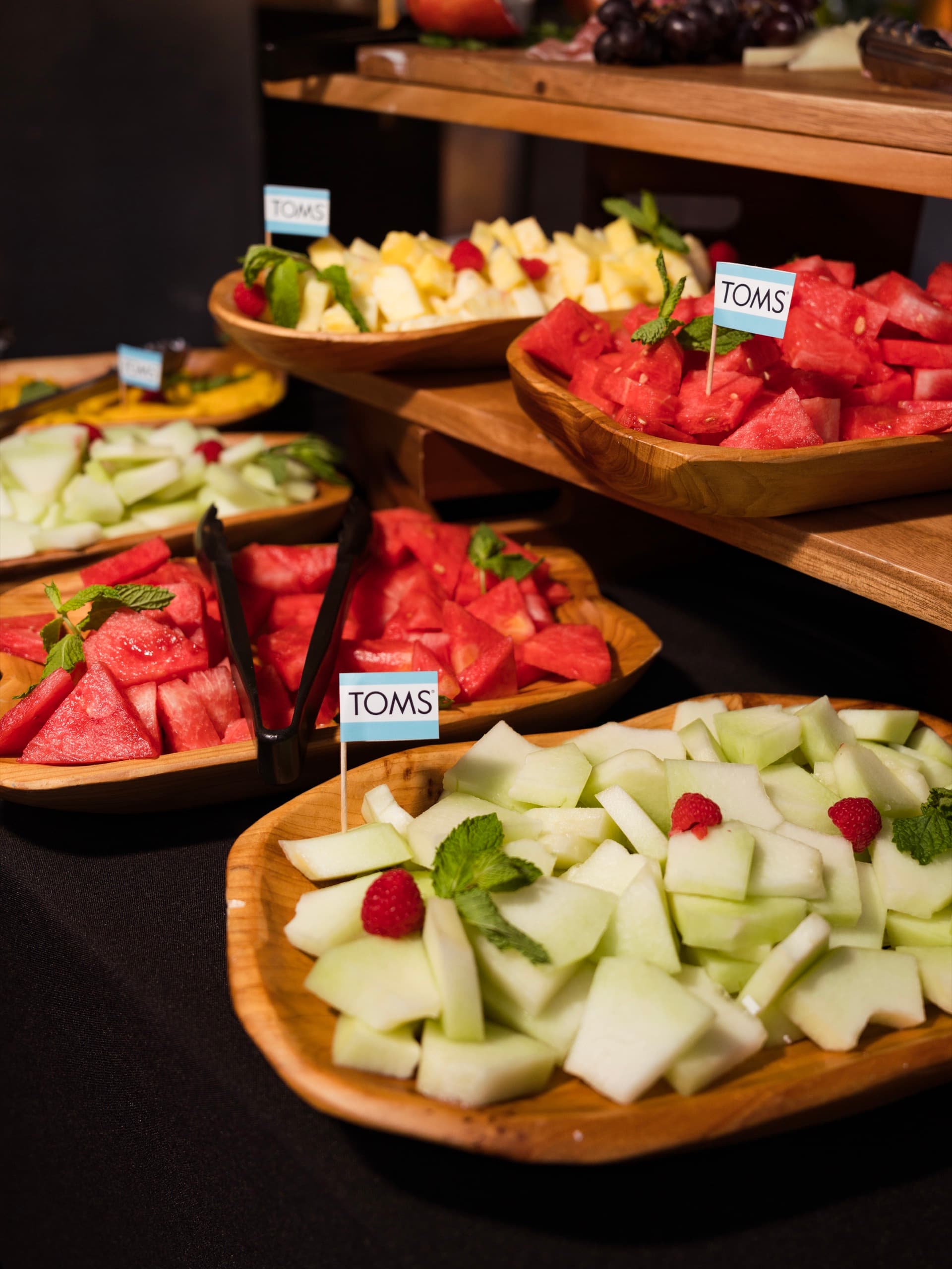 Carved wooden bowls of watermelon, honeydew and pineapple with mint and raspberries, flagged with TOMS cards.