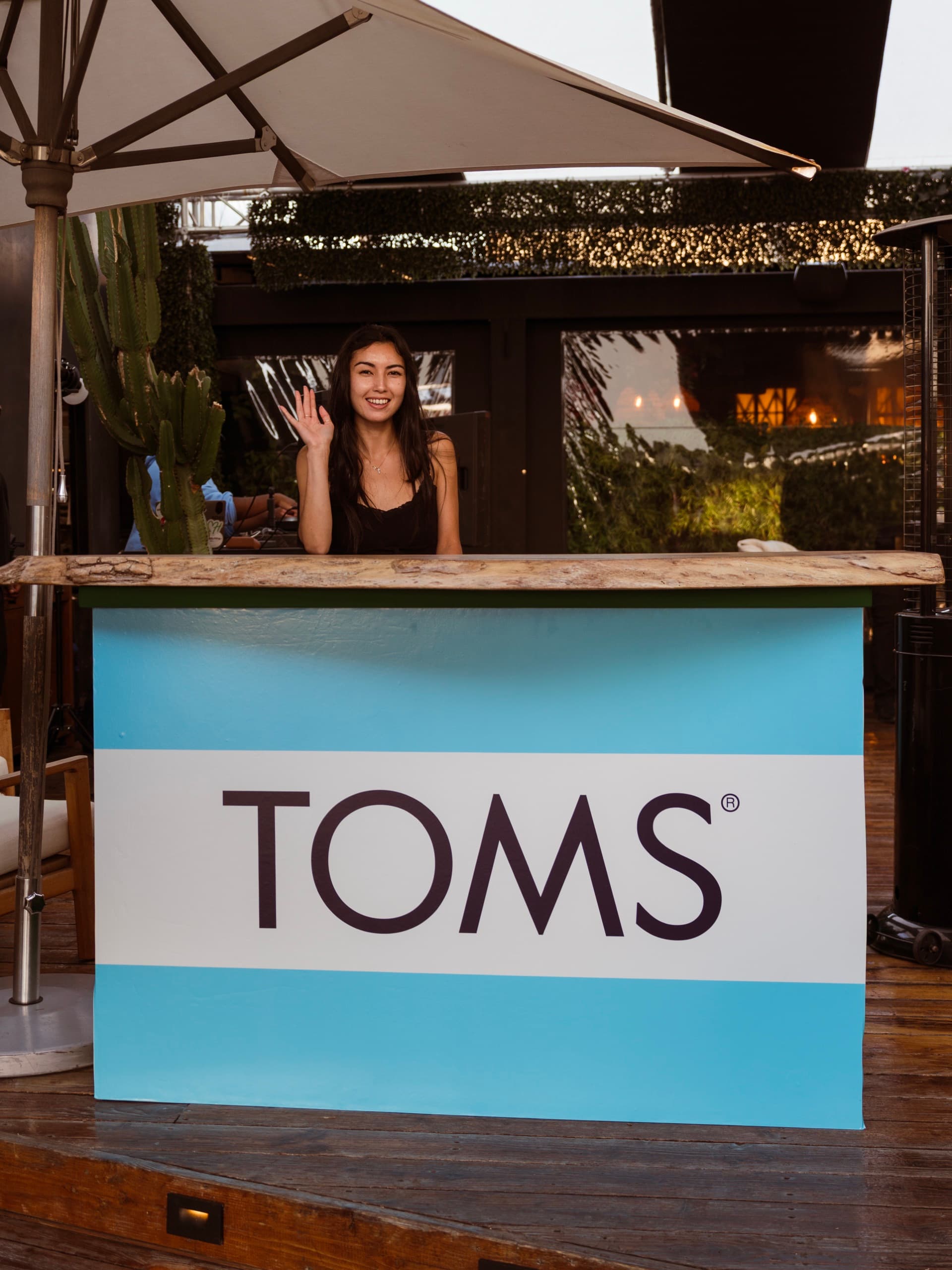 A host waving from behind the TOMS-branded bar at Paloma, under a canvas umbrella beside a cactus.