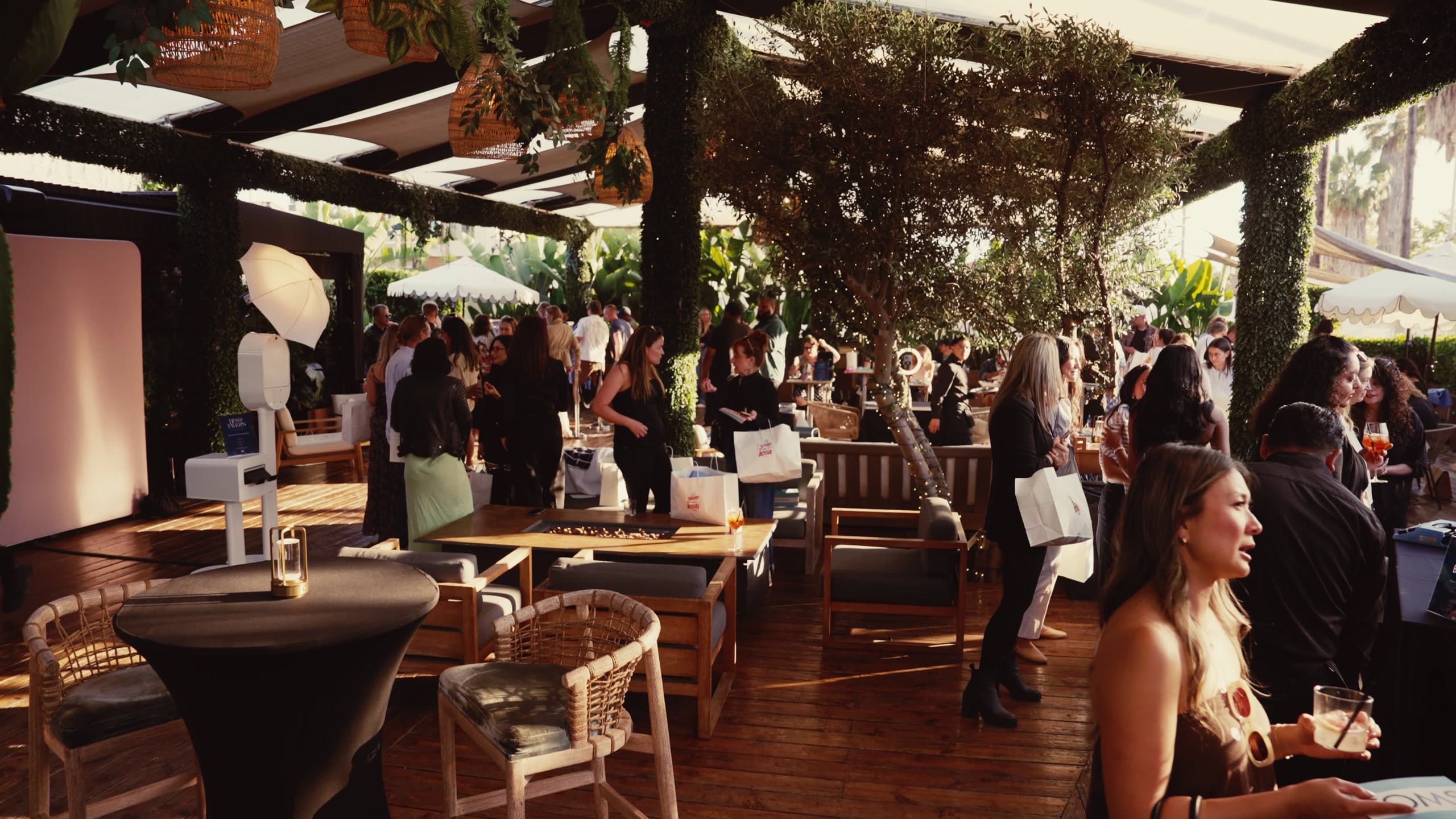 Guests filling the covered terrace at Paloma in late afternoon light, under a pergola hung with olive branches and rattan lanterns, with drinks and TOMS shopping bags in hand.